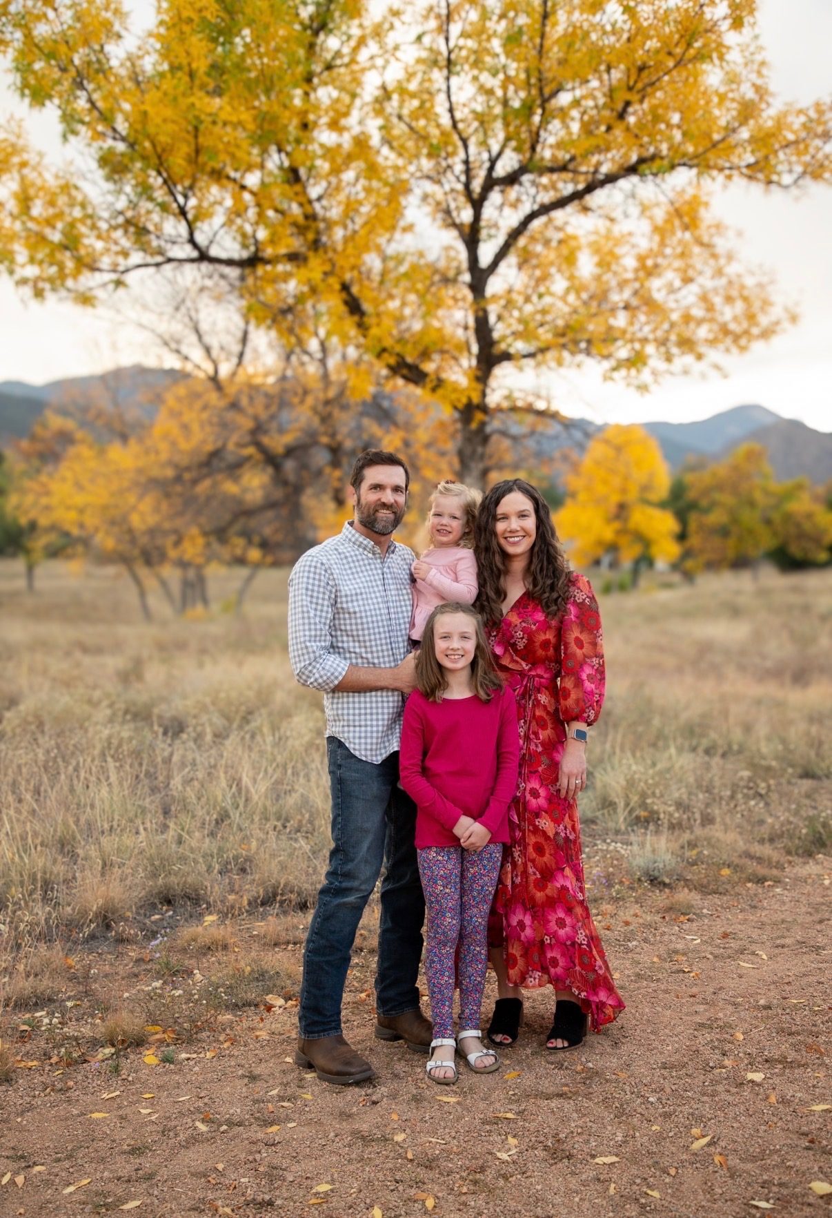 A family of four standing together in a meadow beneath a golden aspen, mountains behind them.