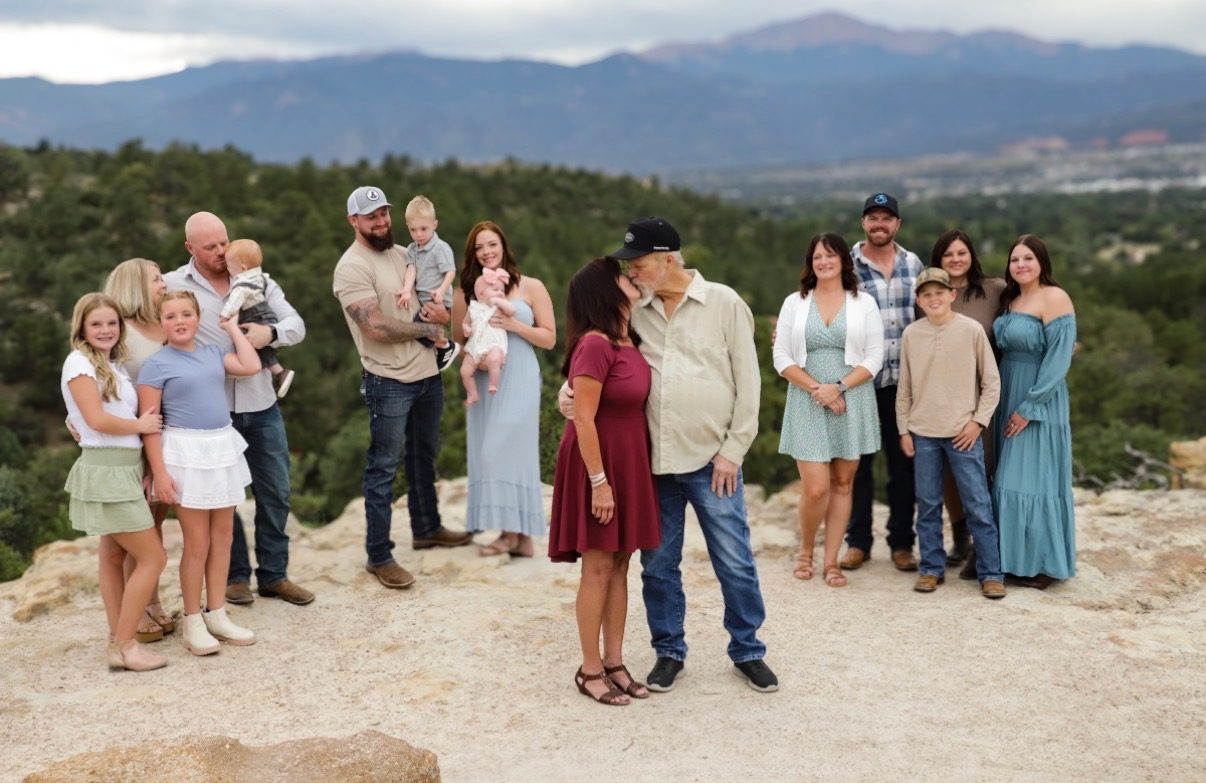A large extended family gathered on a rock overlook with Pikes Peak in the distance, grandparents kissing at the centre.