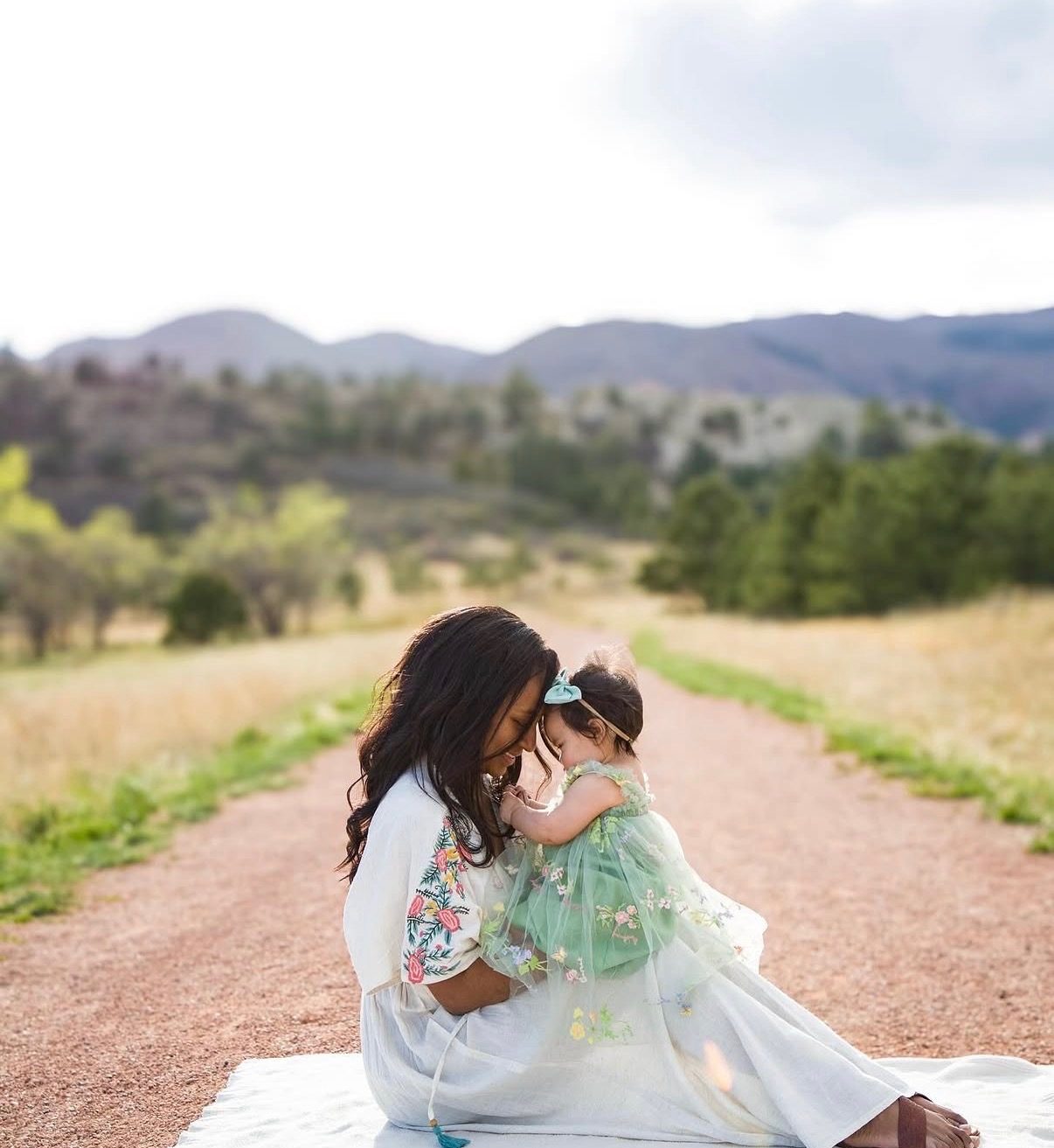 A mother kneeling on a path holding her toddler daughter, foreheads touching, foothills behind them.
