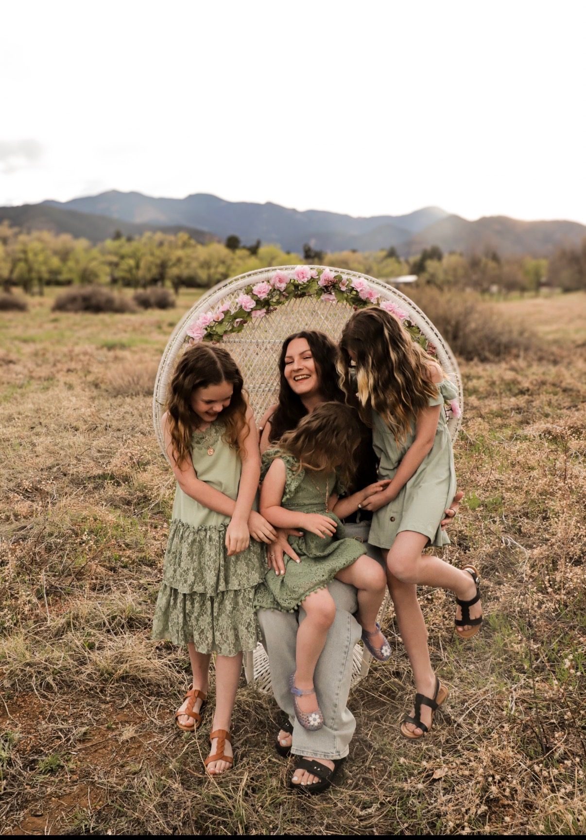 A mother laughing in a wicker chair in a meadow while her three daughters climb around her.