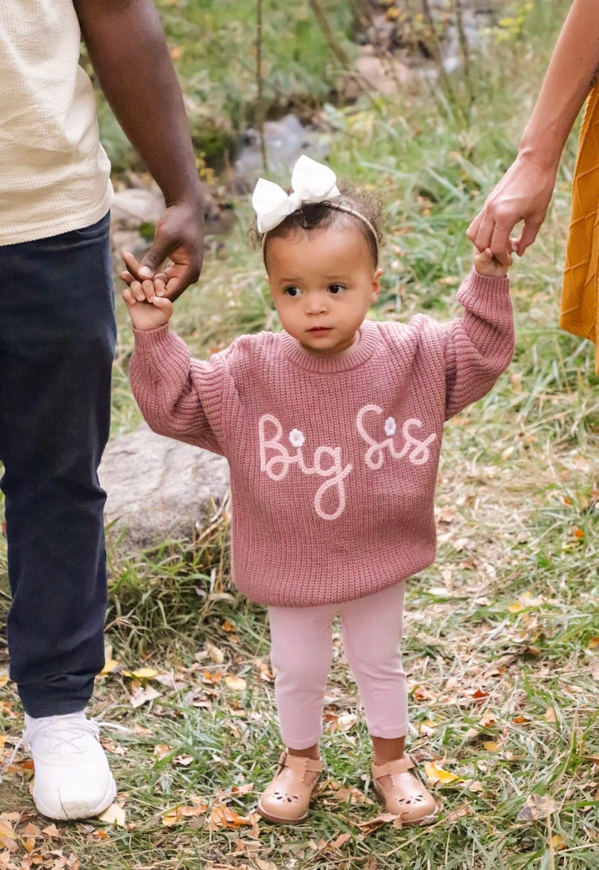 A toddler in a pink sweater reading Big Sis, holding both parents' hands and looking at the camera.