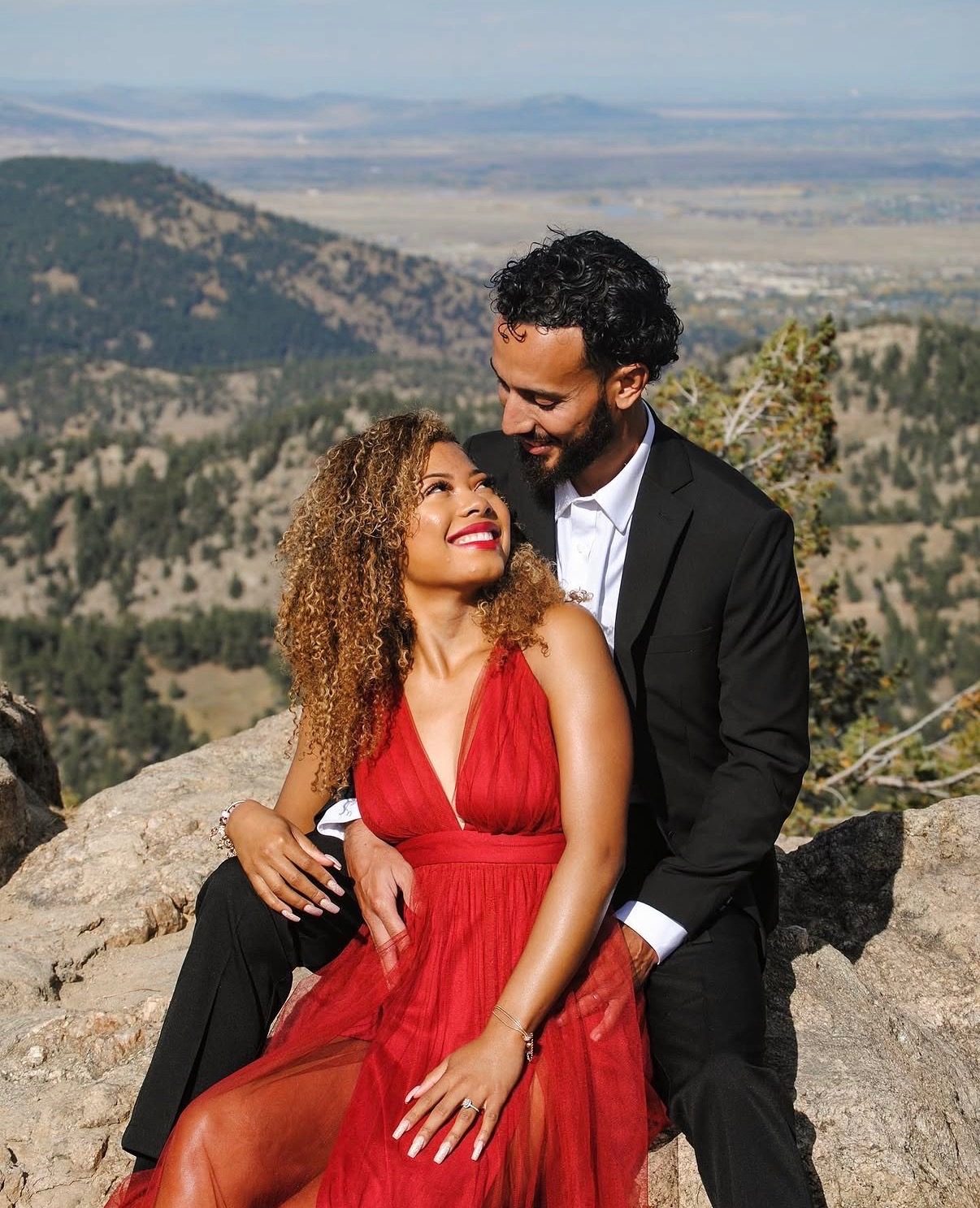An engaged couple sitting on a rock outcrop above the Front Range foothills, she in a red gown looking up at him, he in a black suit.
