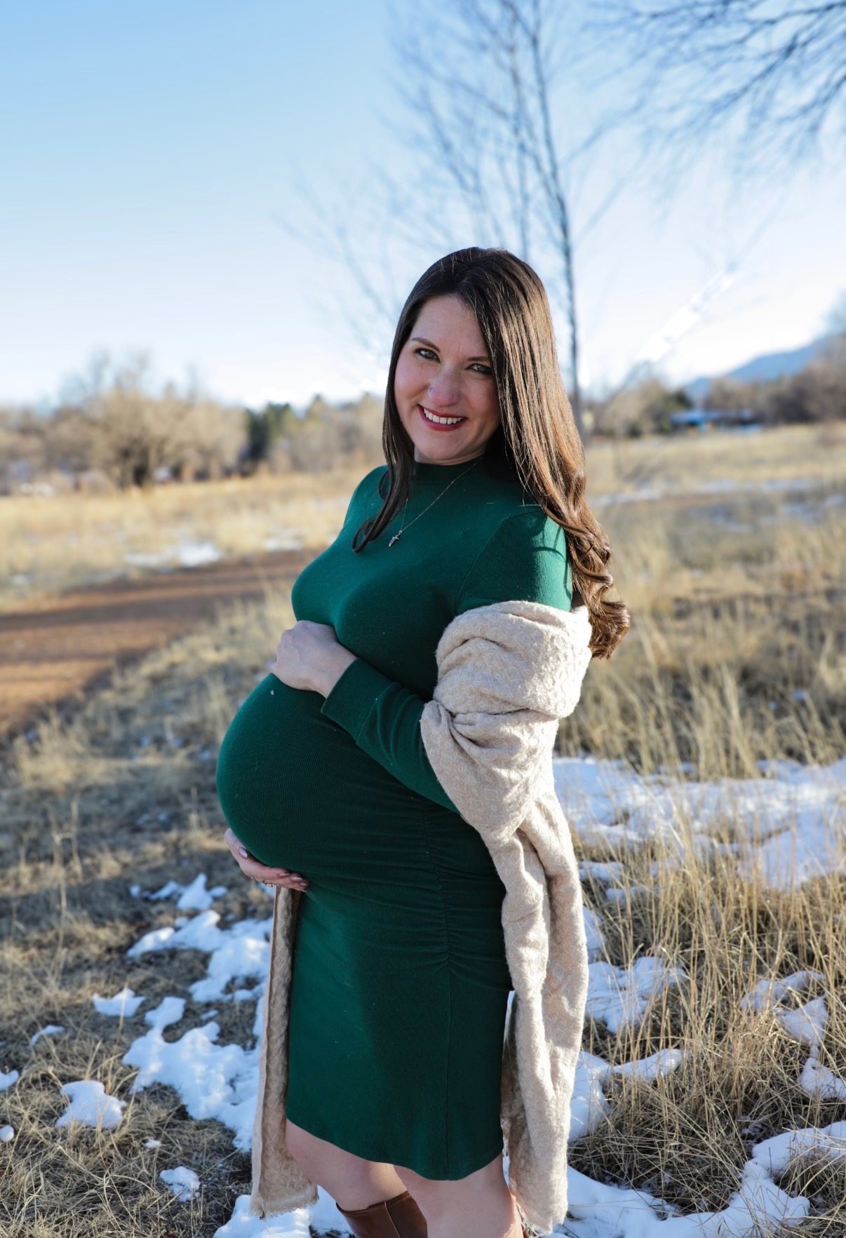 An expectant mother in a green dress standing in a snowy field, holding her bump and smiling.