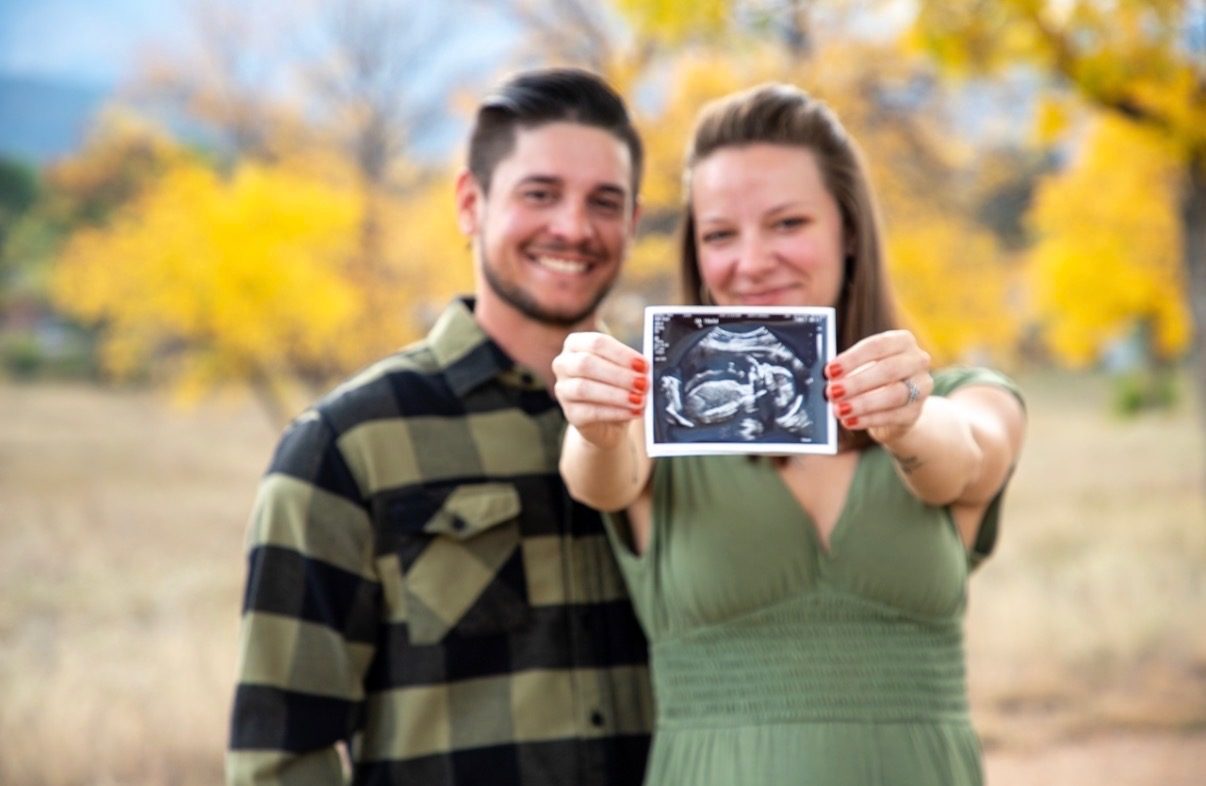 A couple in a field of autumn trees holding up an ultrasound photo toward the camera.