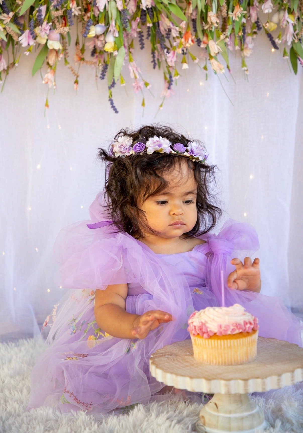 A one-year-old in a lilac tulle dress reaching toward a cupcake beneath hanging flowers.