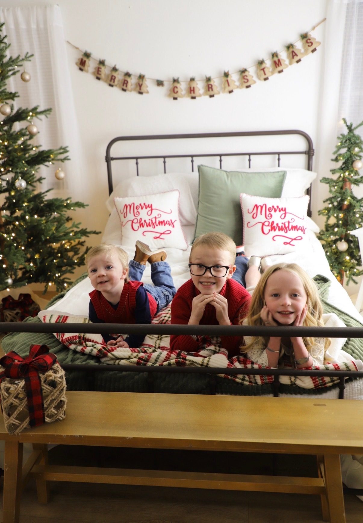 Three children lying on a bed in front of a Merry Christmas garland, chins in their hands.
