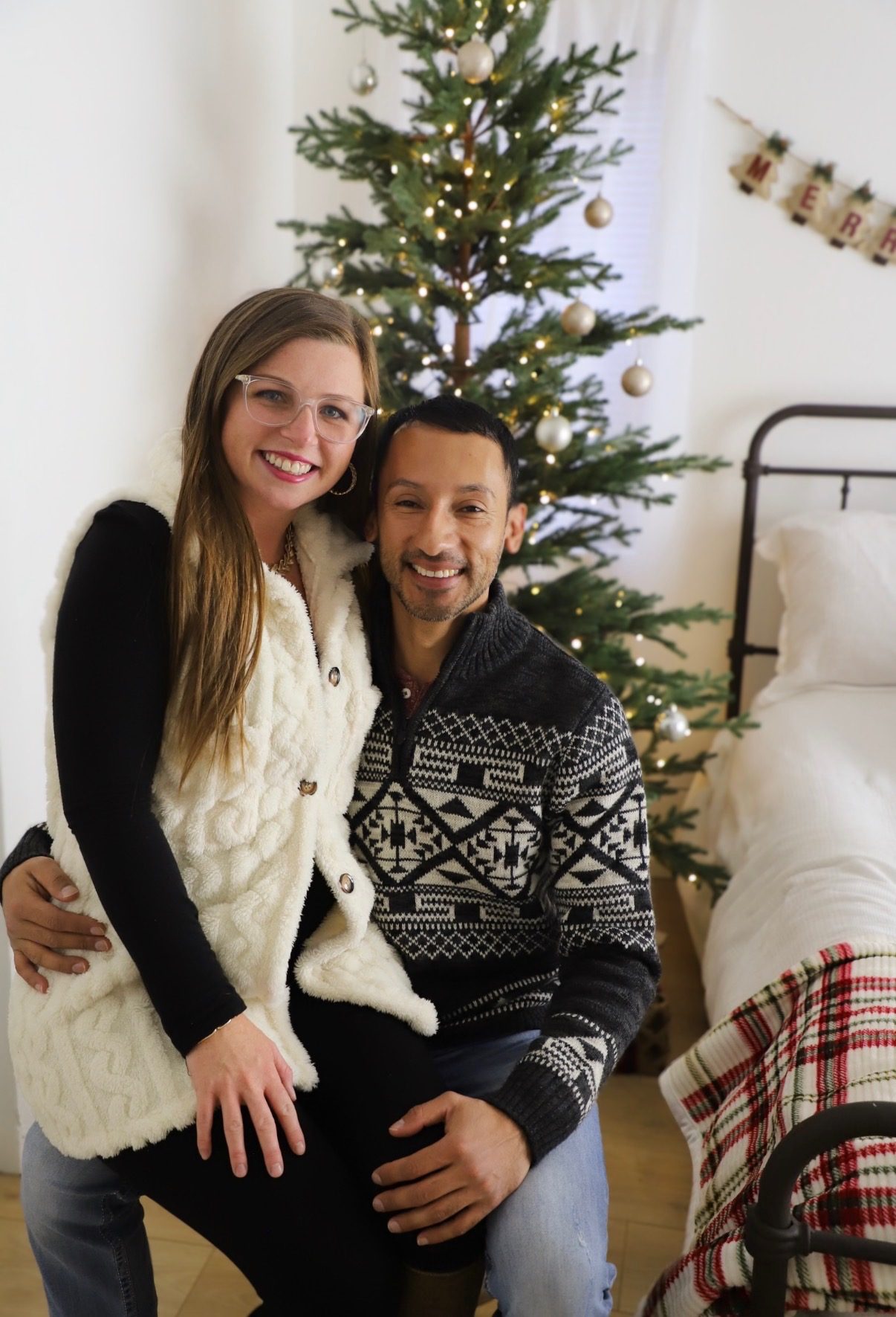 A couple sitting together in front of a lit Christmas tree in a bright studio.