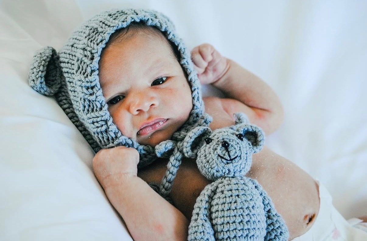 A newborn in a knitted bear bonnet lying beside a small crocheted teddy.