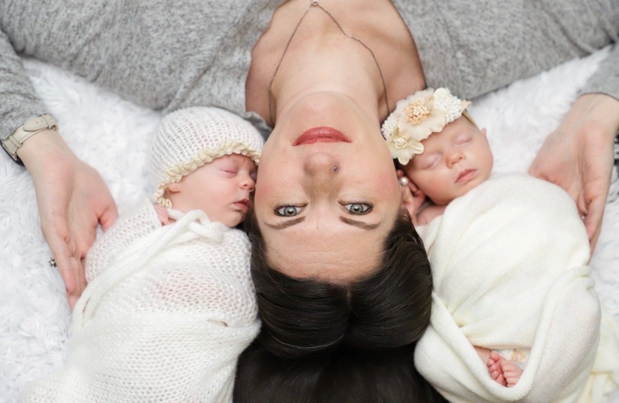 An overhead photograph of a mother lying down with a swaddled newborn asleep on either side of her head.