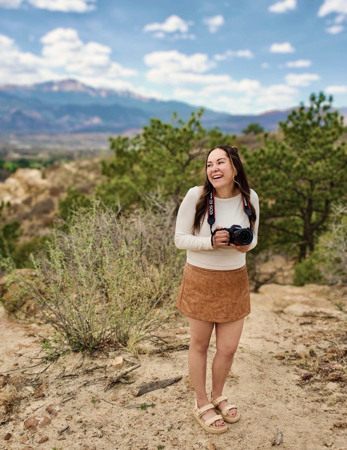 Nicole standing on a trail with her camera, laughing, with Pikes Peak and the Front Range behind her.